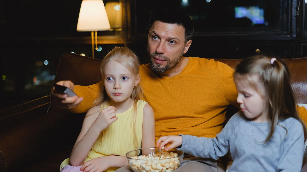 Pai e duas filhas no sofá, comendo pipoca durante uma noite de filme em casa