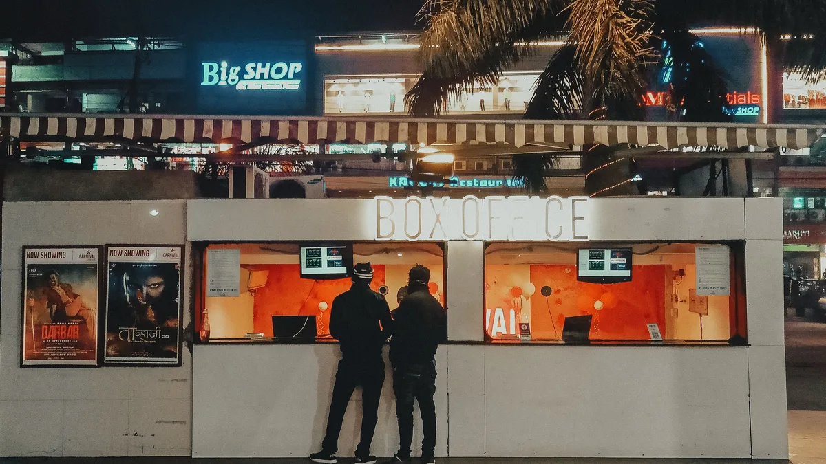 Duas pessoas compram ingressos em uma bilheteria de cinema iluminada por néon à noite