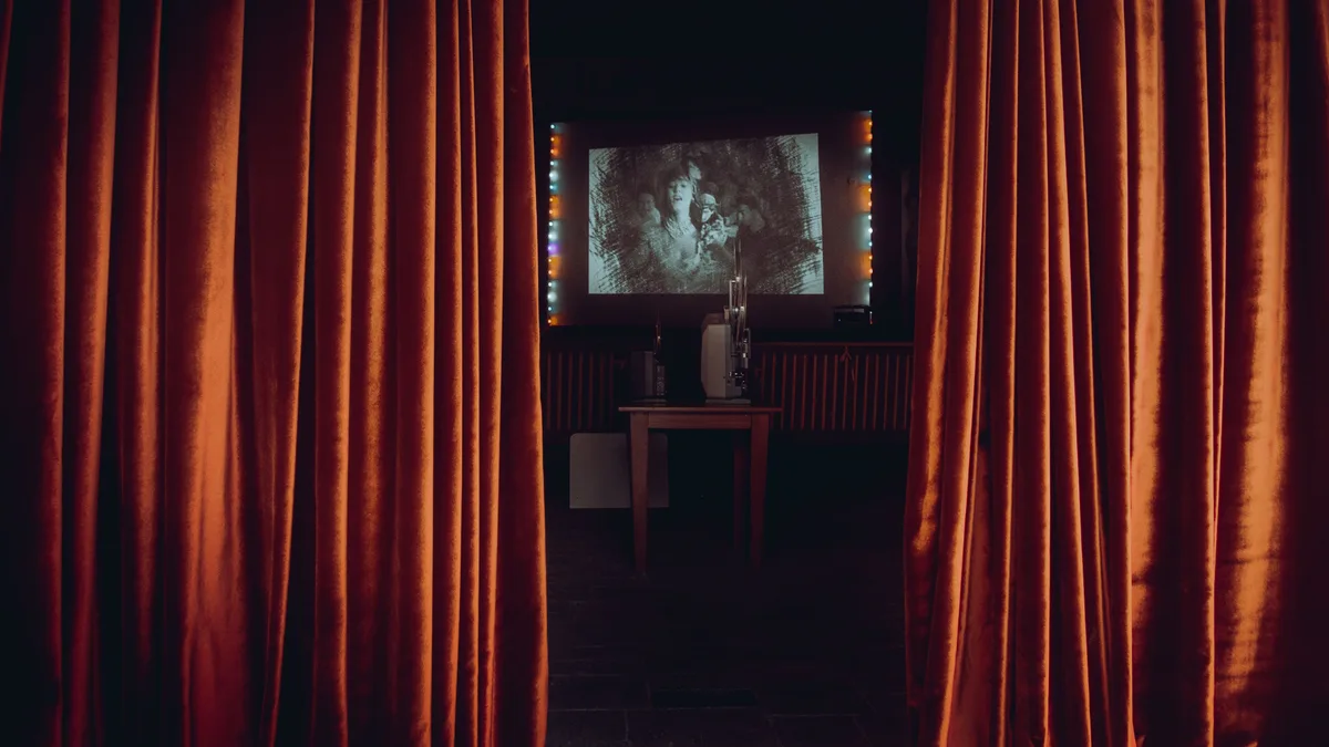 Sala de cinema vista por trás das cortinas vermelhas, com filme clássico projetado na tela