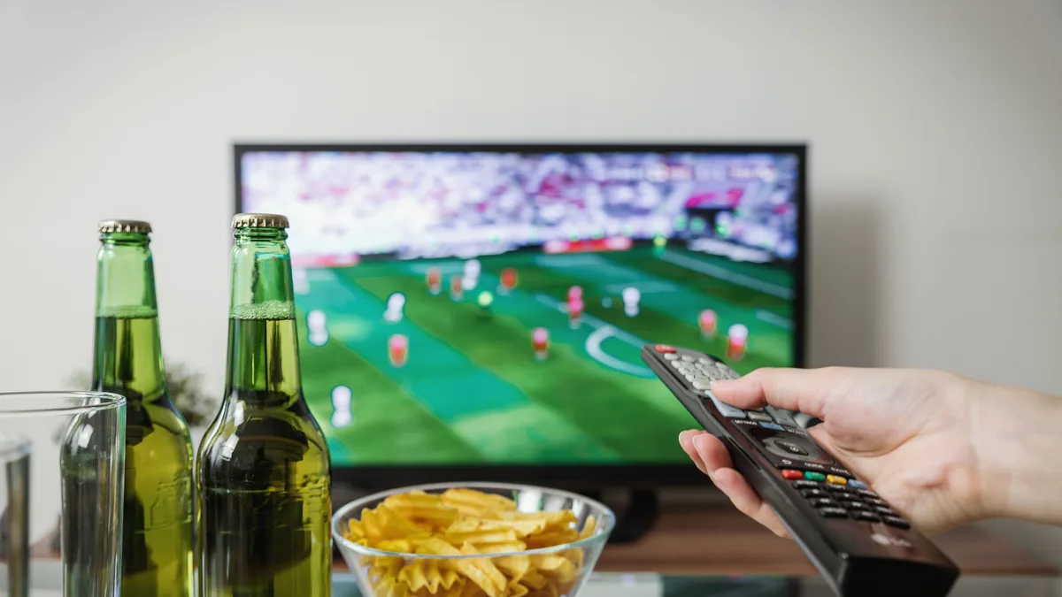 Pessoa segura controle remoto diante de TV que exibe partida de futebol, com petiscos na mesa