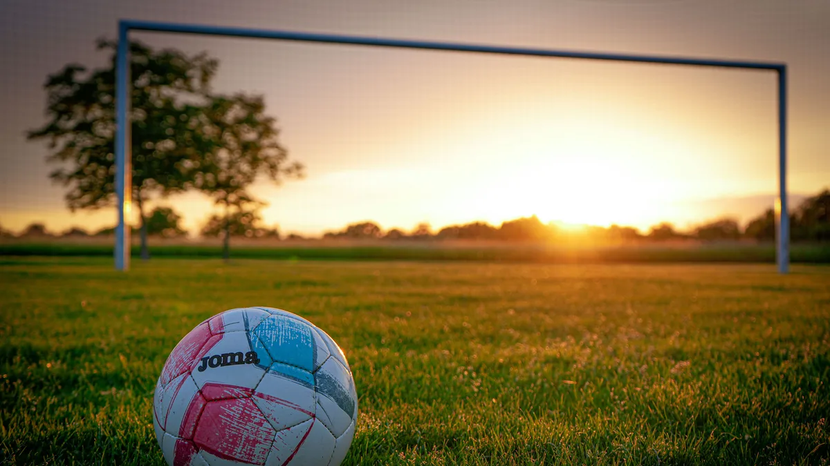 Bola de futebol no gramado diante da trave, iluminada pelo pôr do sol